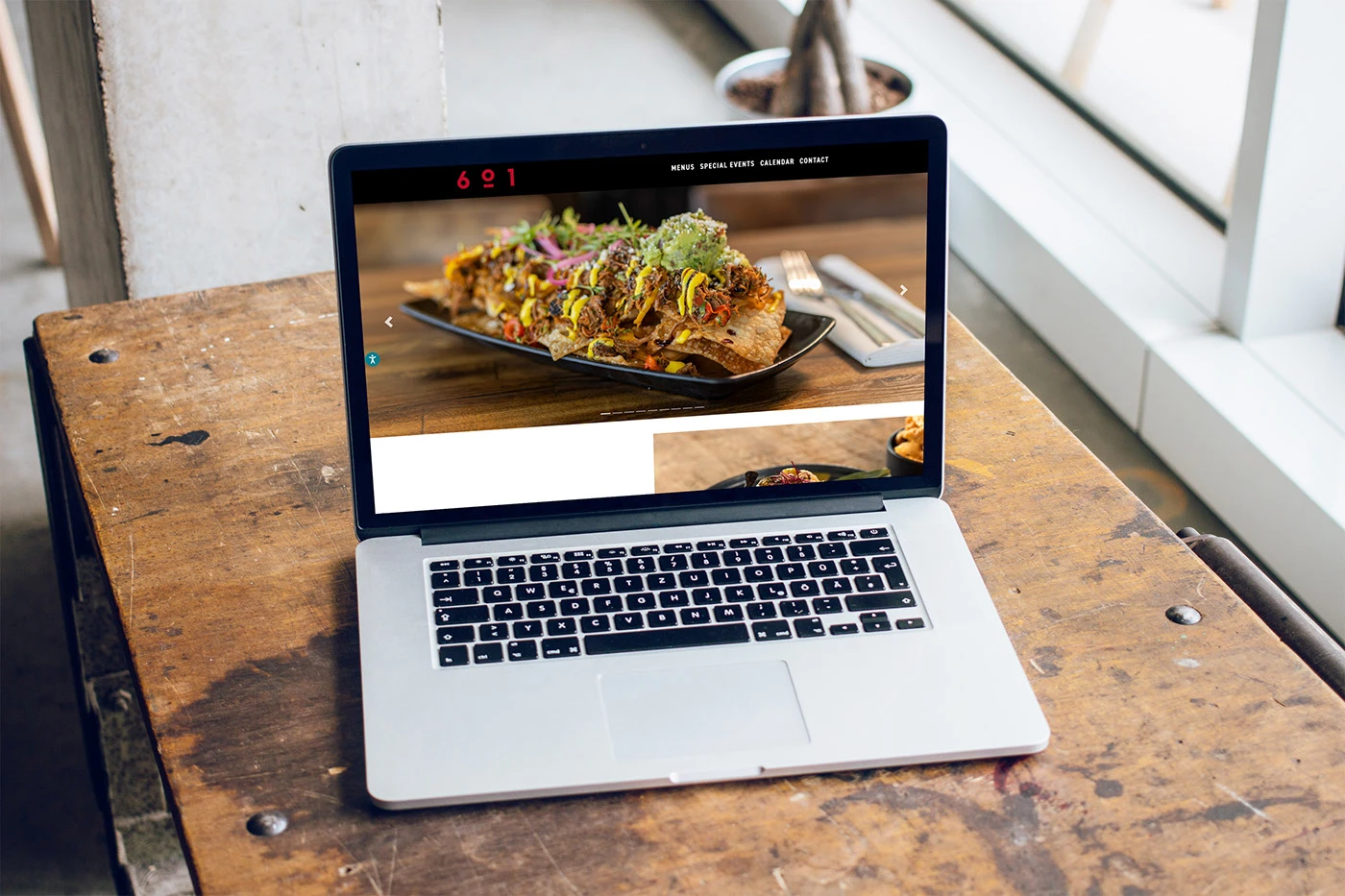 The 601 Miami homepage open on a laptop resting on a worn wooden table, showing the full width food photograph and the 601 navigation.